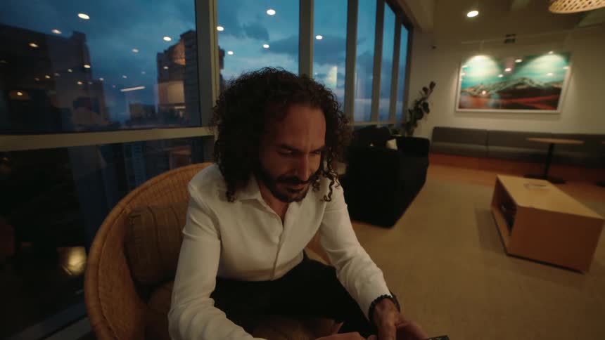 A focused businessman with long curly hair sits in a modern office lounge, scrolling on his smartphone as evening falls over the city outside the window