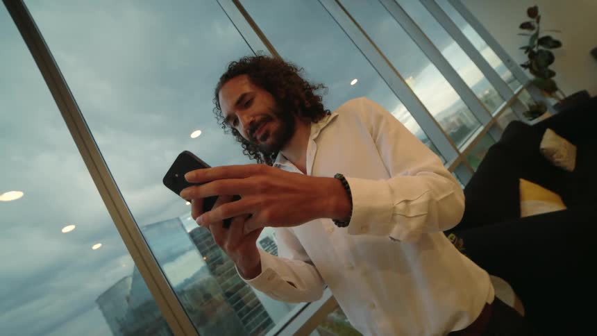 A businessman with long curly hair smiles while using his smartphone in a modern high-rise office overlooking the city
