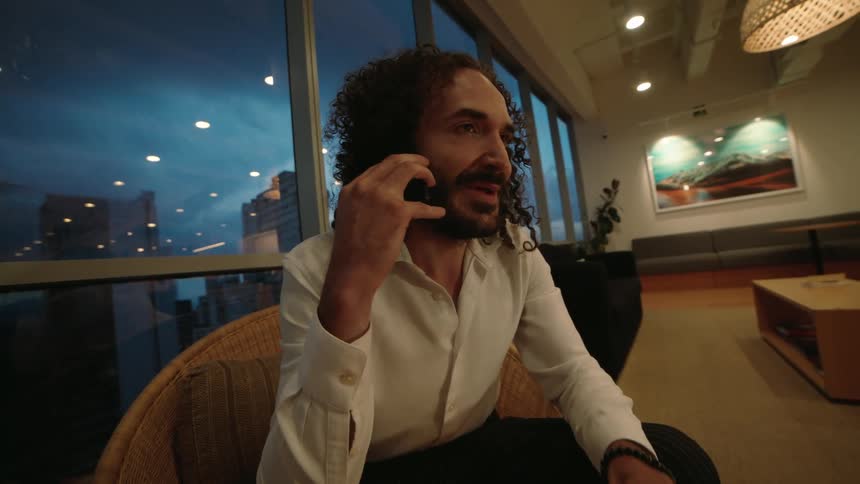 A stylish businessman with curly hair gestures while on a phone call in a modern high-rise office at dusk