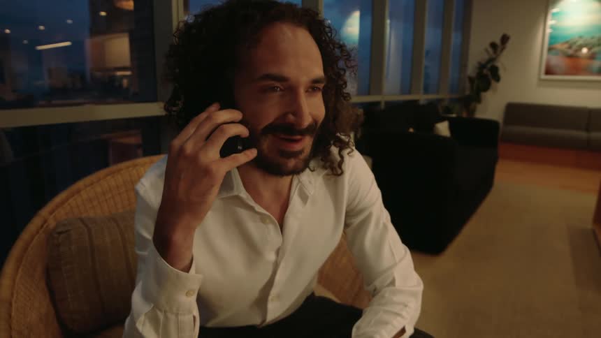 A smiling man with long curly hair answers a pleasant phone call in his stylish living room in the evening