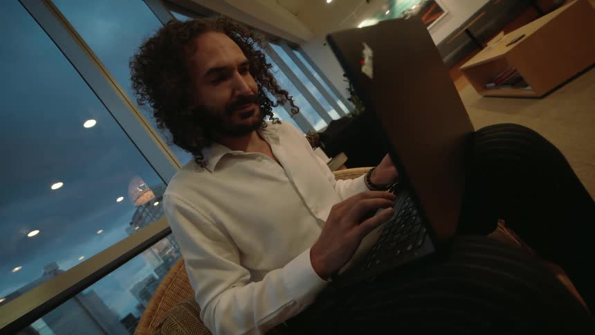 A professional man with a beard and curly hair works on a laptop in a stylish lounge as evening falls over the city outside the window