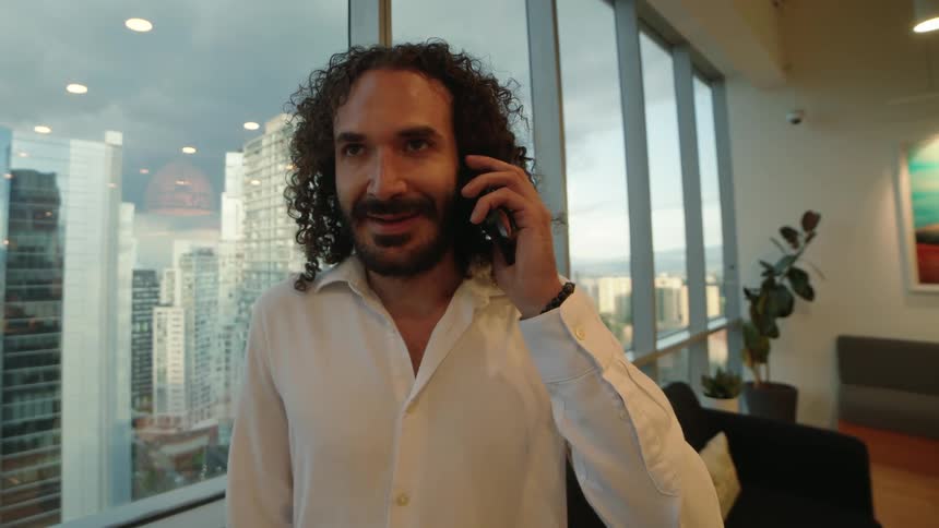 Smiling businessman with curly hair answers a call on his smartphone in a high-rise office overlooking the city