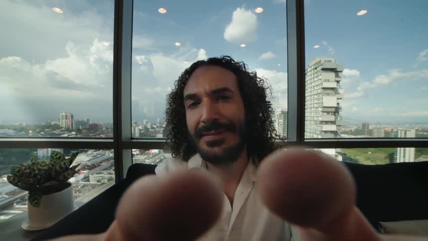 POV of a man with long curly hair looking directly into the camera from a high-rise apartment overlooking a cityscape