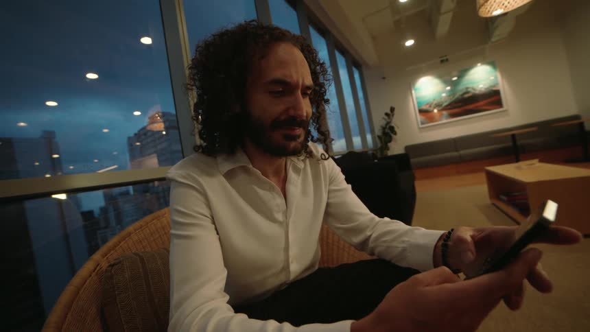 A stylish man with curly hair sits in a modern lounge at dusk, scrolling and texting on his smartphone with a city skyline view