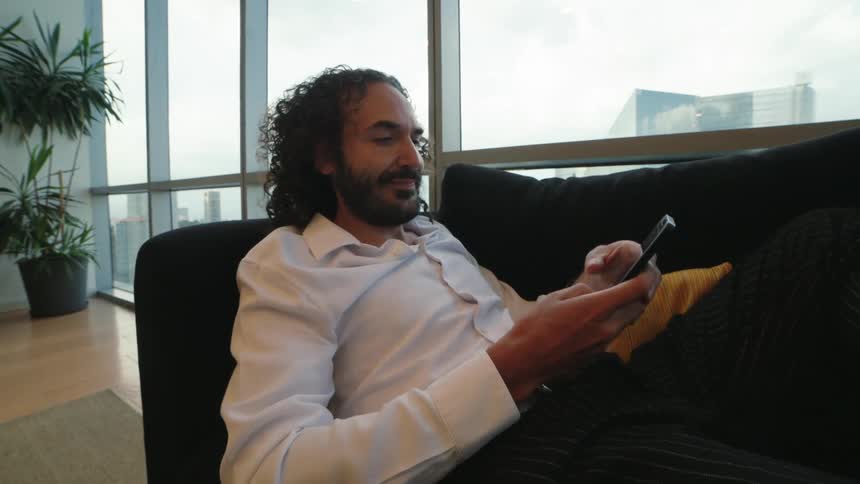 A stylish man with long curly hair and a beard ends a phone call and smiles while texting on his smartphone, lounging on a sofa in a modern penthouse