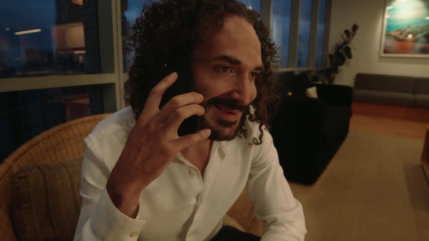 A handsome man with curly hair smiles while talking on his phone in his modern apartment at dusk