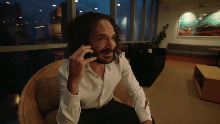 A handsome, smiling businessman with curly hair has a pleasant conversation on his phone in a modern office with a city view at night