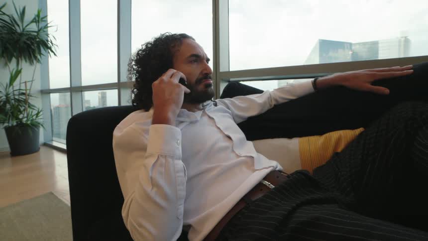 A relaxed businessman with long curly hair talks on his phone while lounging on a couch in a modern high-rise apartment
