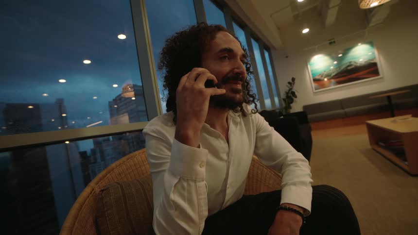 A businessman with curly hair makes a phone call while sitting in a modern high-rise office lounge at night