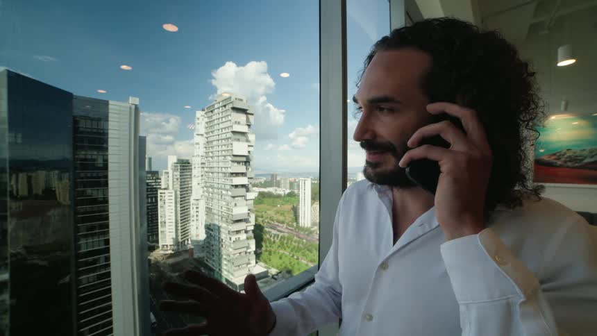 A happy businessman with long hair talks on his phone while looking out the window of a high-rise office overlooking the city
