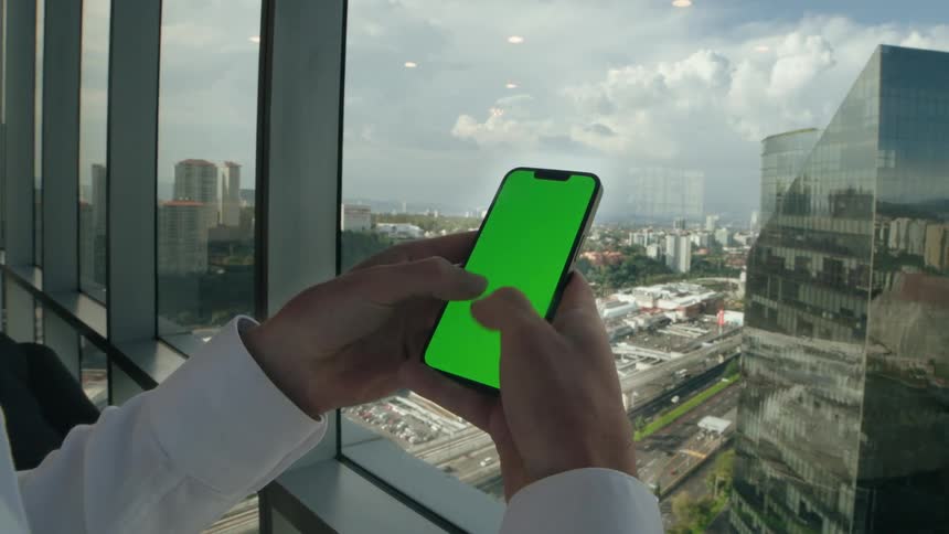 A businessman in a white shirt uses a green screen smartphone while standing by a window in a high-rise office overlooking the city