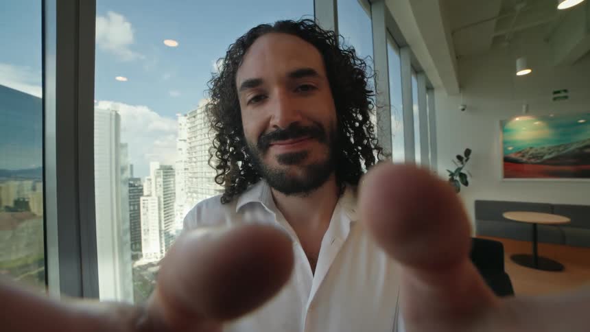 A man with long curly hair holds the camera for a selfie-style video call in a modern office with a city view