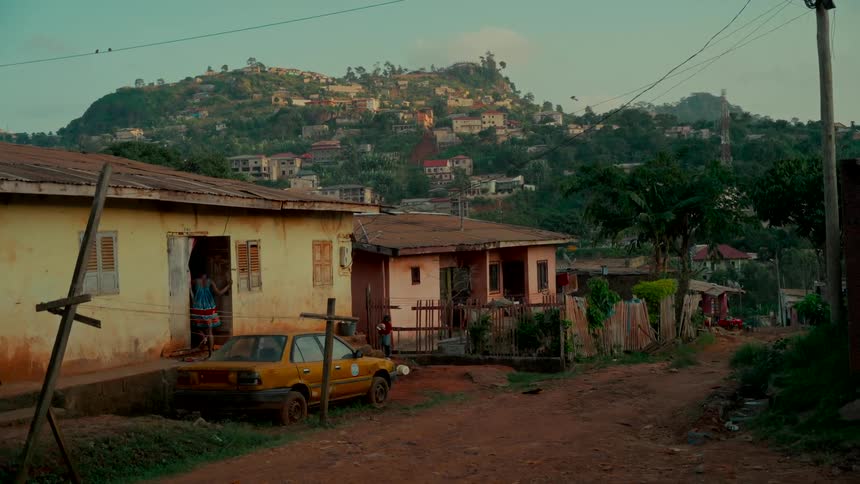A wide, static shot of a tranquil village in Sierra Leone, with homes scattered across a lush hill and a woman standing in her doorway