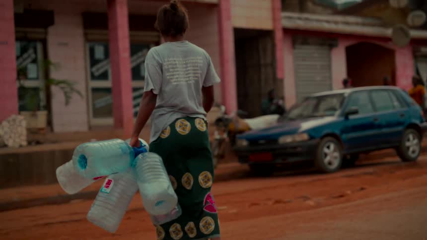 Slow motion following shot of a person carrying a large bundle of empty plastic water bottles down a dusty village street