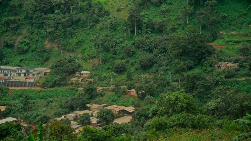 A red vehicle drives up a winding dirt road through a rural village nestled in a lush, green, forested valley