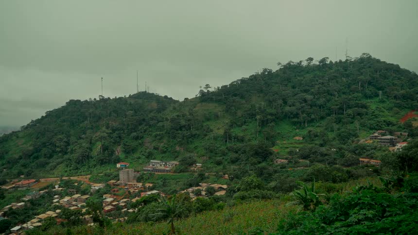 A static wide shot of a lush green mountain range with communication towers and a village nestled at its base under a cloudy sky