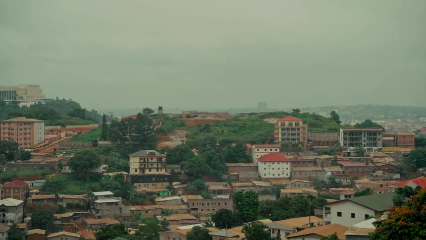 A slow pan reveals a sprawling cityscape with diverse buildings nestled among lush green hills on a foggy, overcast day