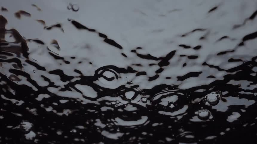 A hypnotic, worm's-eye view of raindrops creating abstract ripples on a dark water surface in slow motion