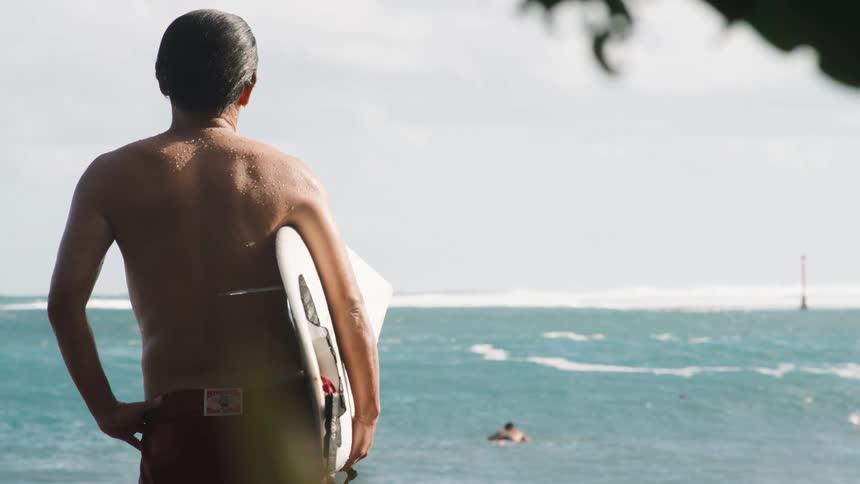 A surfer with his back to the camera holds his surfboard while looking out at the ocean waves in slow motion