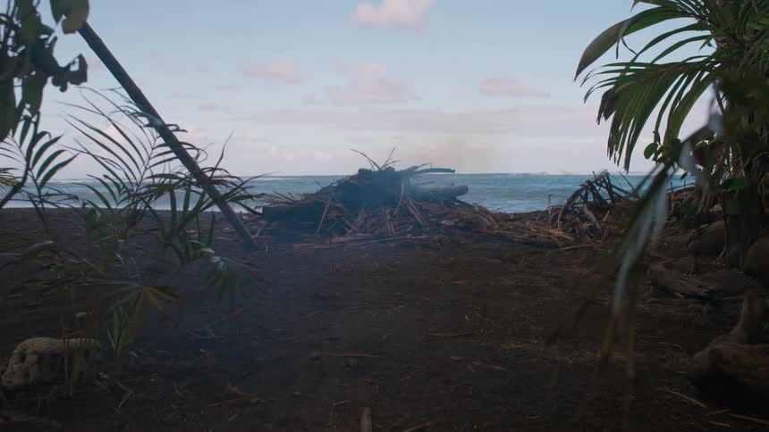 Smoke rises from a smoldering campfire on a dark, sandy beach with the ocean in the background