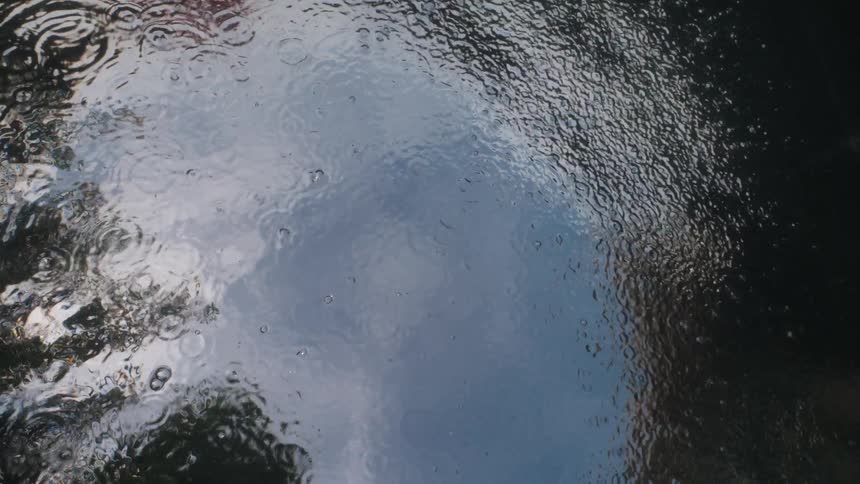 A top-down view of raindrops creating ripples on the surface of water, reflecting the overcast sky