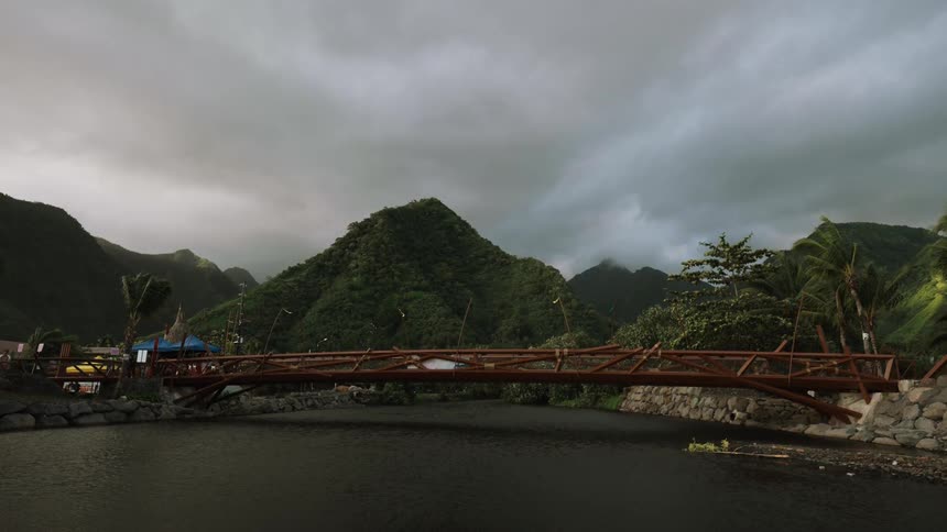 A wooden bridge spans a dark river in a lush tropical valley under a moody, overcast sky
