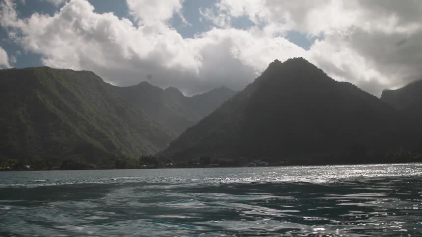 Dramatic clouds and sunlight backlight a lush green tropical mountain range as seen from the tranquil ocean water