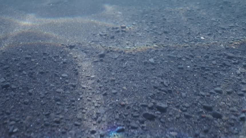 An immersive journey underwater reveals dancing light and rainbow caustics on the dark sandy bottom