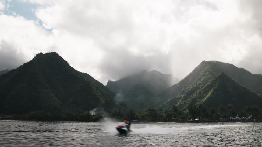 A person on a jet ski races across the water in slow motion with a backdrop of dramatic, lush green mountains on a tropical island