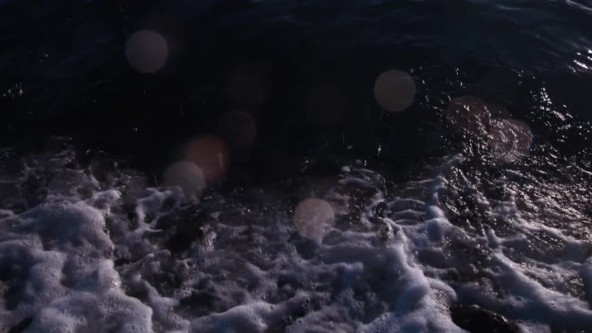 A dramatic slow-motion shot captures a dark ocean wave breaking over smooth shoreline rocks
