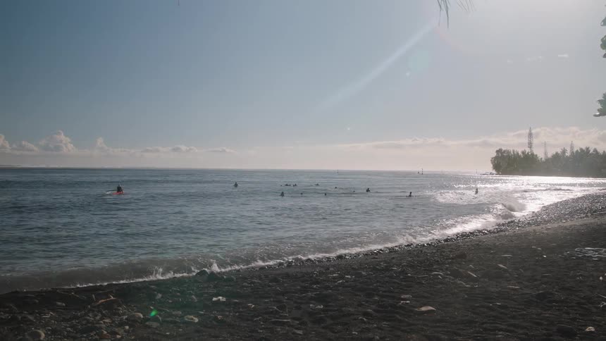 A scenic view from a dark sand beach shows surfers waiting for waves in the calm ocean on a sunny day