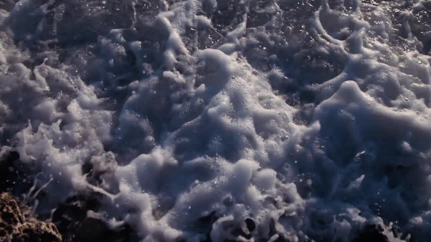Foamy white ocean waves wash over dark glistening rocks in a super slow-motion close-up