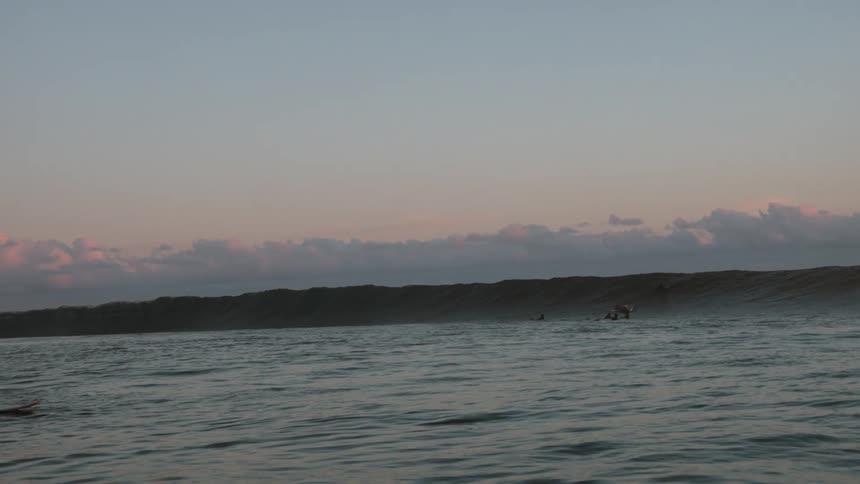 A group of surfers wait patiently in the ocean as a long, powerful wave begins to break at dusk
