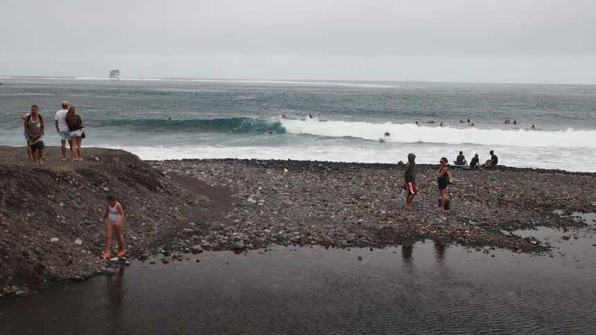 Spectators watch from a rocky shore as surfers ride waves in the ocean on a cloudy day, while a young girl plays by a calm inlet