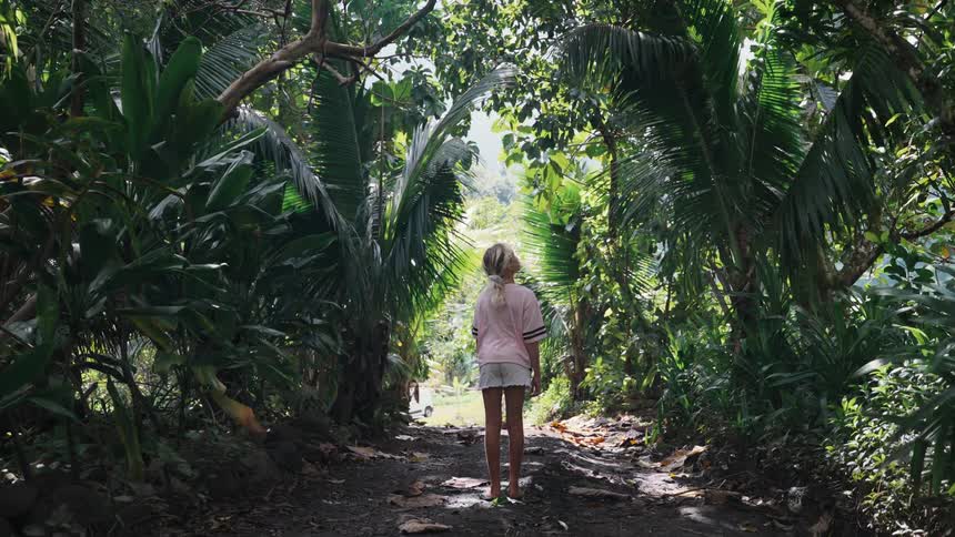 A young blonde girl stands on a pathway surrounded by a lush tropical jungle