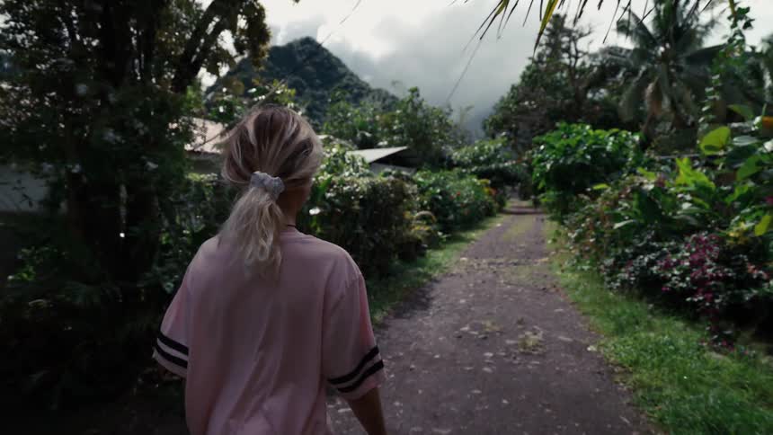 Slow motion shot from behind a woman with a blonde ponytail as she walks down a path through a lush tropical village