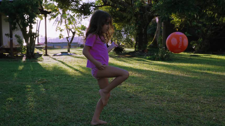 A young girl in a pink shirt kicks an orange ball into the air in slow motion while playing on a grassy lawn