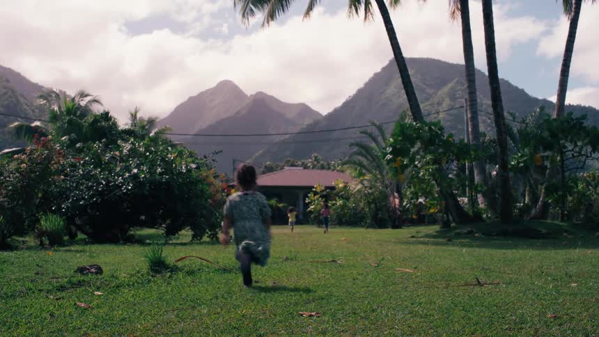 Three young children joyfully run across a lush green lawn towards a house in a stunning tropical setting with mountains in the background