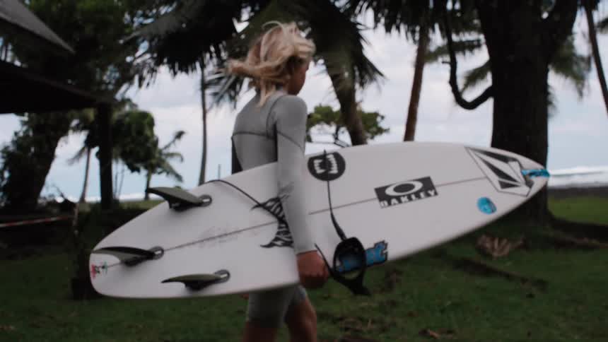 Side-view slow-motion of a young surfer in a wetsuit carrying a surfboard towards a tropical beach with palm trees