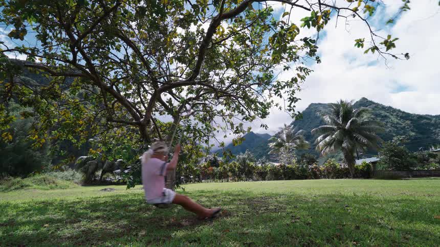 A carefree blonde woman enjoys a slow motion swing under a tree in a lush tropical paradise with mountains in the background