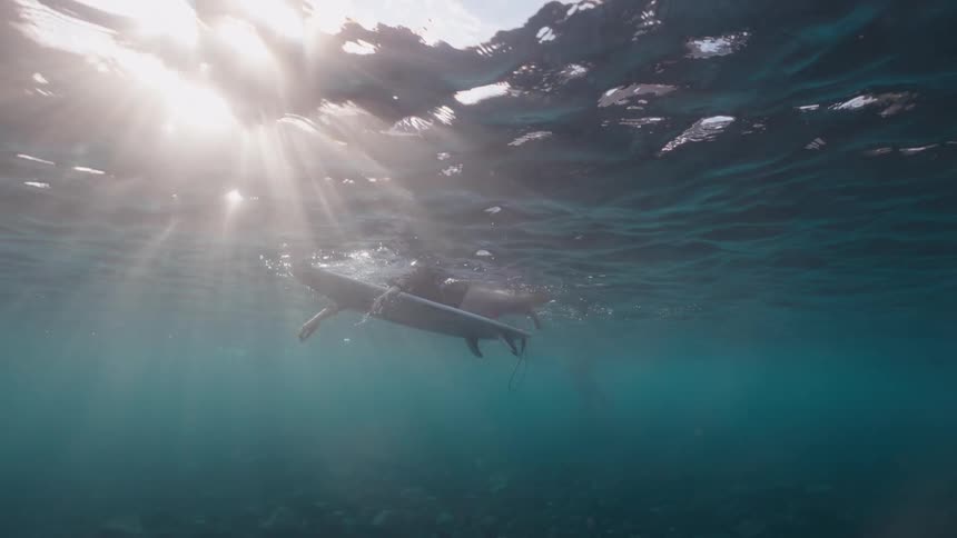 An underwater view shows a surfer floating on their board as sun rays pierce through the tranquil ocean surface