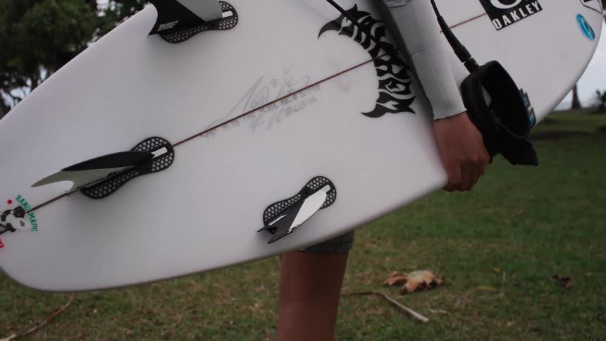 Slow-motion close-up of a surfer in a wetsuit carrying their white surfboard across a grassy area