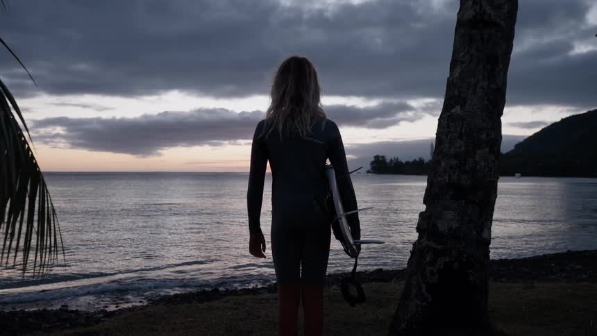 A young surfer in a wetsuit stands with their board on a tropical beach, looking out at the ocean as the sun sets under a cloudy sky
