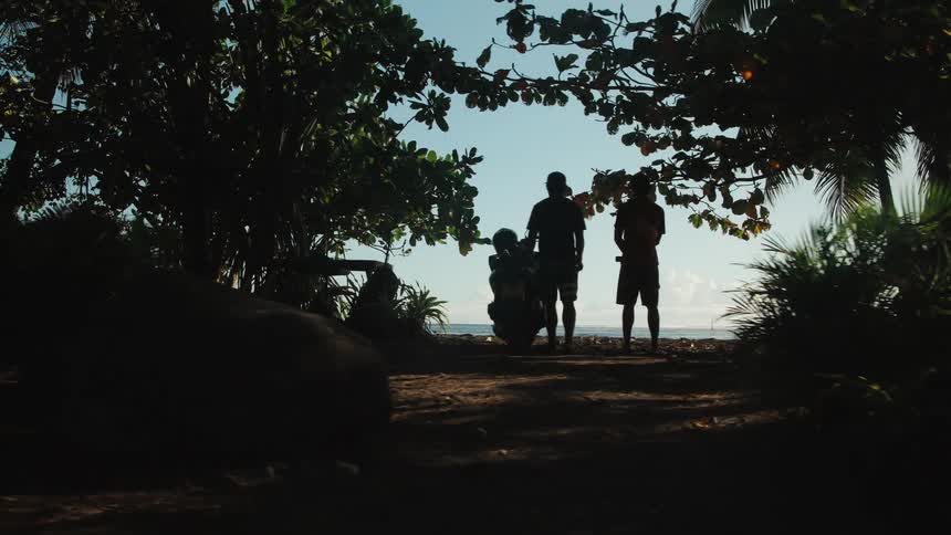 A slow reveal from behind dark tropical foliage shows the silhouettes of friends with a motorcycle admiring the ocean view