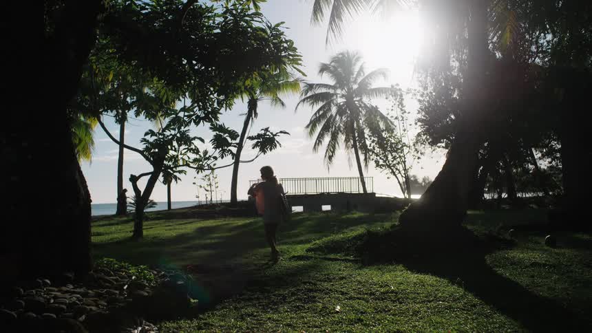 A young female surfer walks through a sun-drenched tropical park with her board, heading towards the ocean