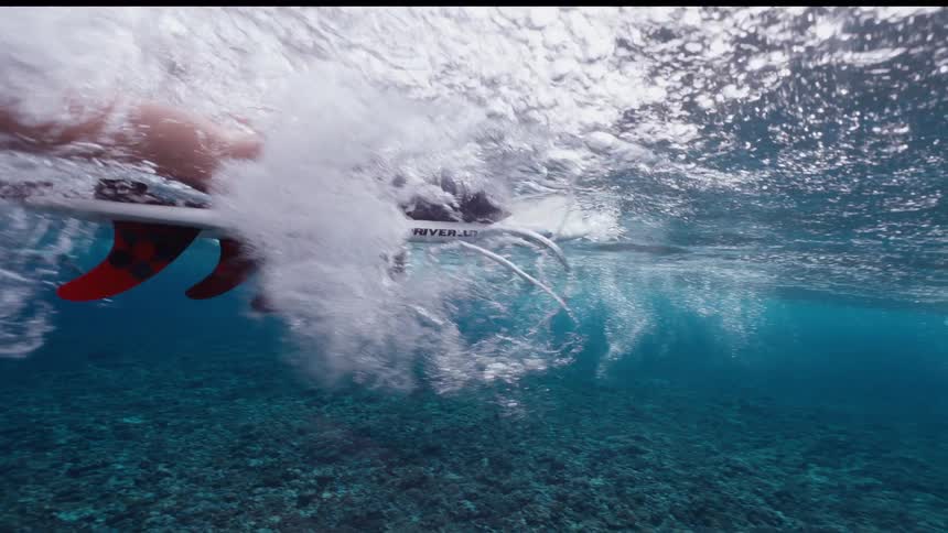 A slow-motion underwater view captures a surfer paddling on their board over a clear blue coral reef