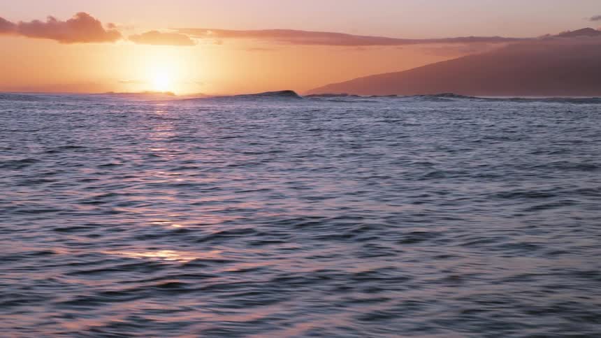 A serene view of the ocean with gentle ripples as the sun sets over the distant mountains, casting a warm golden glow