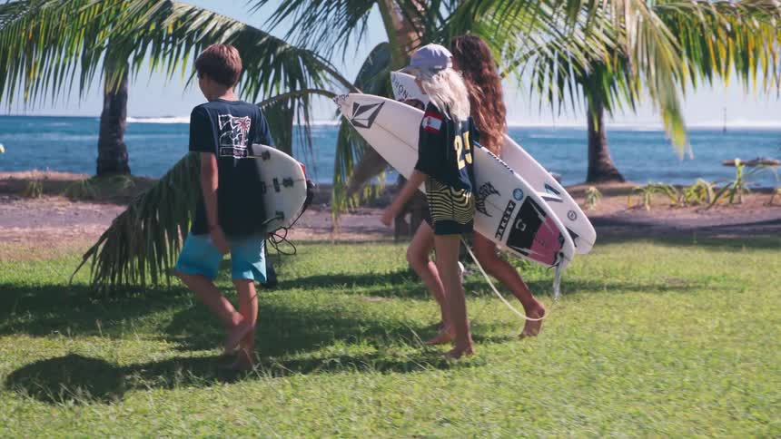 A group of young surfers carry their boards as they walk barefoot across a grassy park towards the tropical ocean