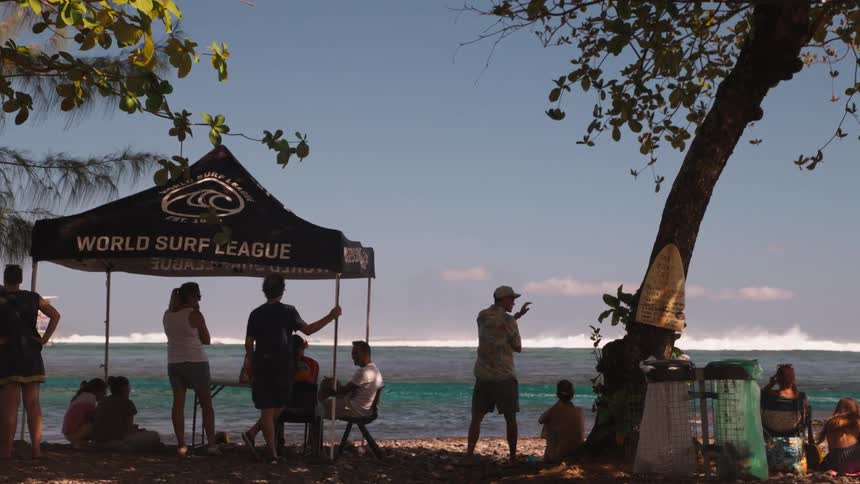 Silhouetted spectators gather under a World Surf League tent on a sunny tropical beach to watch a surfing competition on the ocean waves