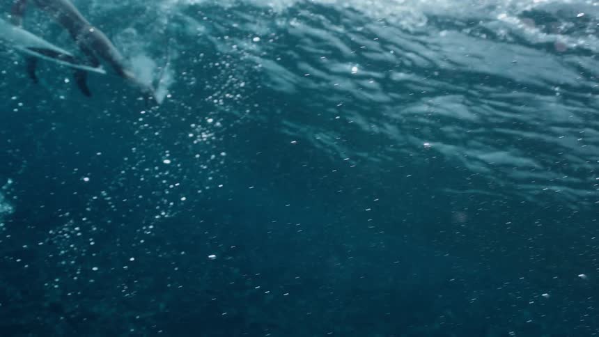 A dramatic underwater slow-motion shot reveals a surfer patiently waiting on their board in the tranquil blue ocean